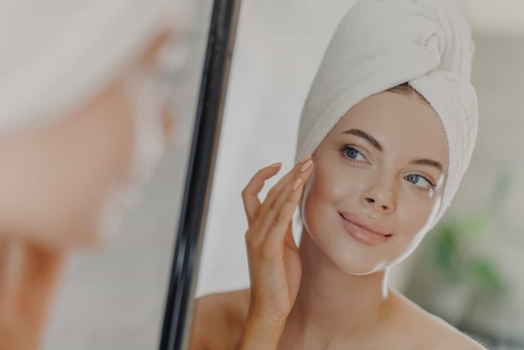 Beautiful woman touches face gently, looks at her reflection in mirror, enjoys softness of skin