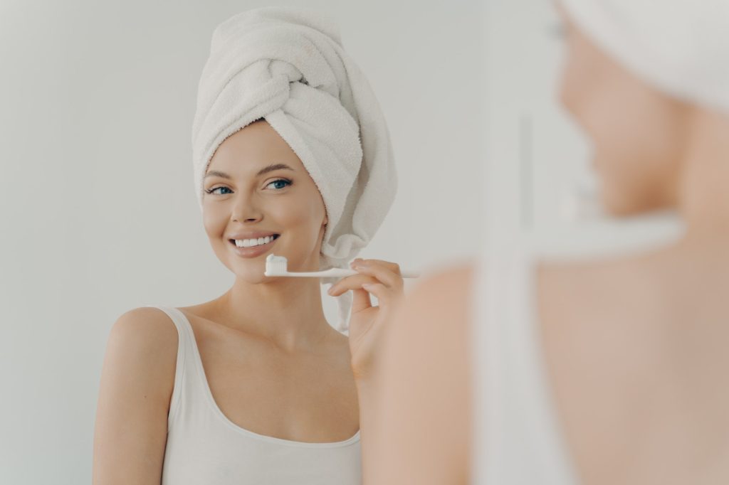 Beautiful young woman with healthy perfect smile brushing teeth and looking in mirror