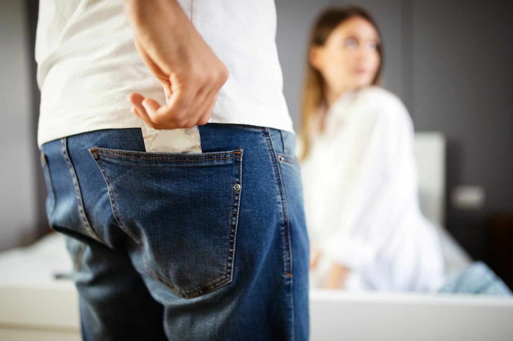 Cropped image of man holding a condom, sexy woman is lying on bed