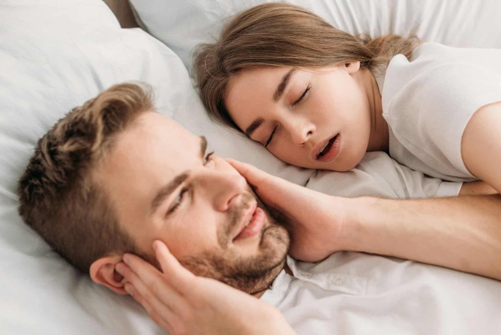 exhausted man plugging ears with hands while lying in bed near snoring wife