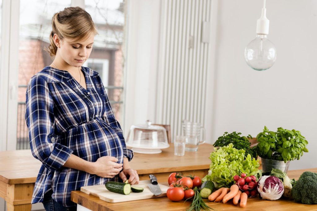 Gravid kvinne som kutter opp grønnsaker. Hun har en rekke flotte og deilige grønnsaker liggende på kjøkkenbenken. Hun har en flott blå stripete skjorte.