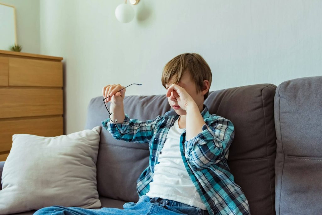 Small school boy rubs his eyes from using glasses for myopia at home. Dry eyes. Pain. Bad eyesight.