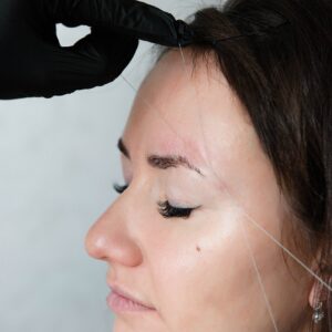 Browist makes eyebrow correction with tweezers thread. A model with dark hair in a light studio is s