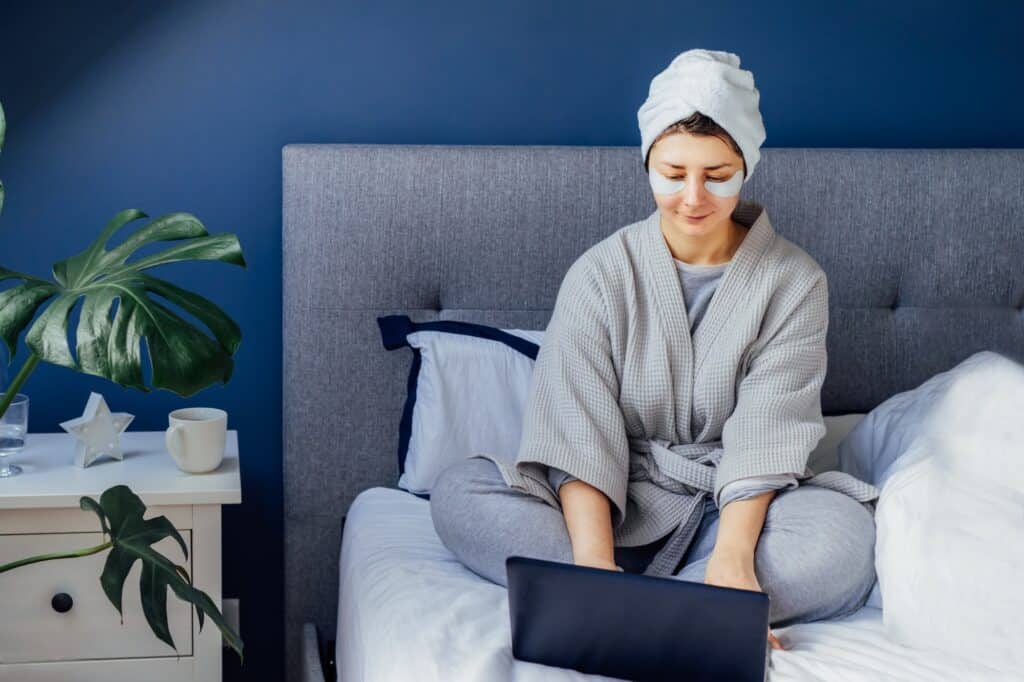 Woman in pajama with eyes patches and towel on head using laptop sitting on her bed. beauty routine