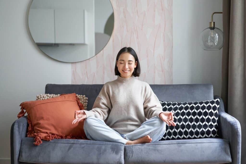 Relaxation and patience. Smiling young asian woman in cozy room, sitting on sofa and meditating