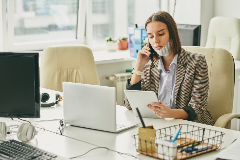 Young pretty brunette office worker with protective mask on chin using tablet