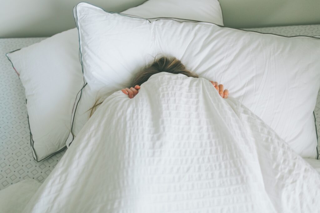 A woman in bed, covering her face with blankets.