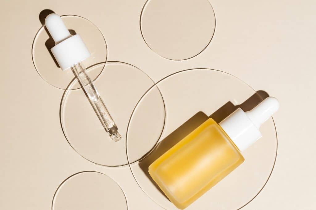 Glass petri dish with bottle dropper of retinol or vit C serum, essential oil on beige background.